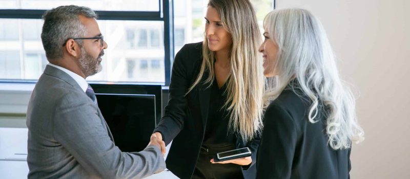 Confident business partners meeting in office, standing and shaking hands, talking, discussing collaboration. Medium shot. Communication or partnership concept